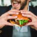 person holding burger bun with vegetables and meat